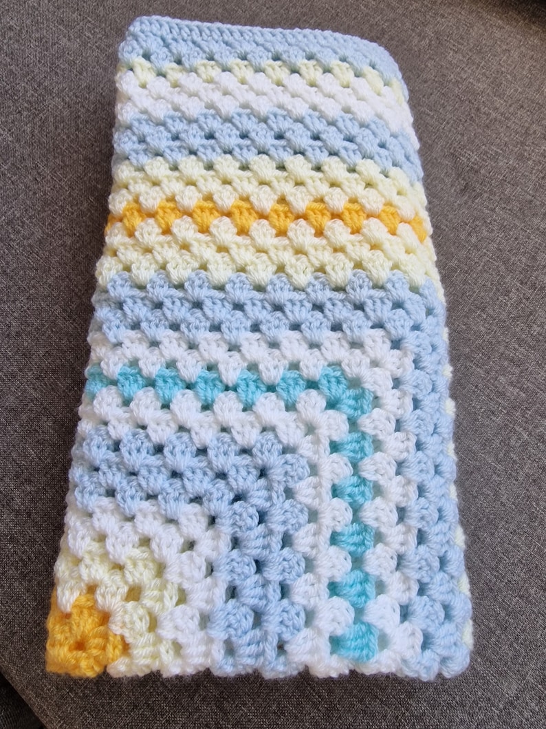 May include: A crocheted baby blanket in shades of blue, white, and yellow. The blanket is made with a granny square pattern and has a diagonal stripe design.