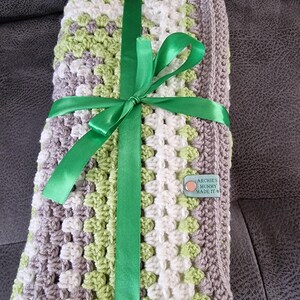 May include: A crocheted baby blanket in shades of gray, white, and green, tied with a green satin ribbon. A small brown tag with the text "Archie's Mummy Made It" is attached to the blanket.