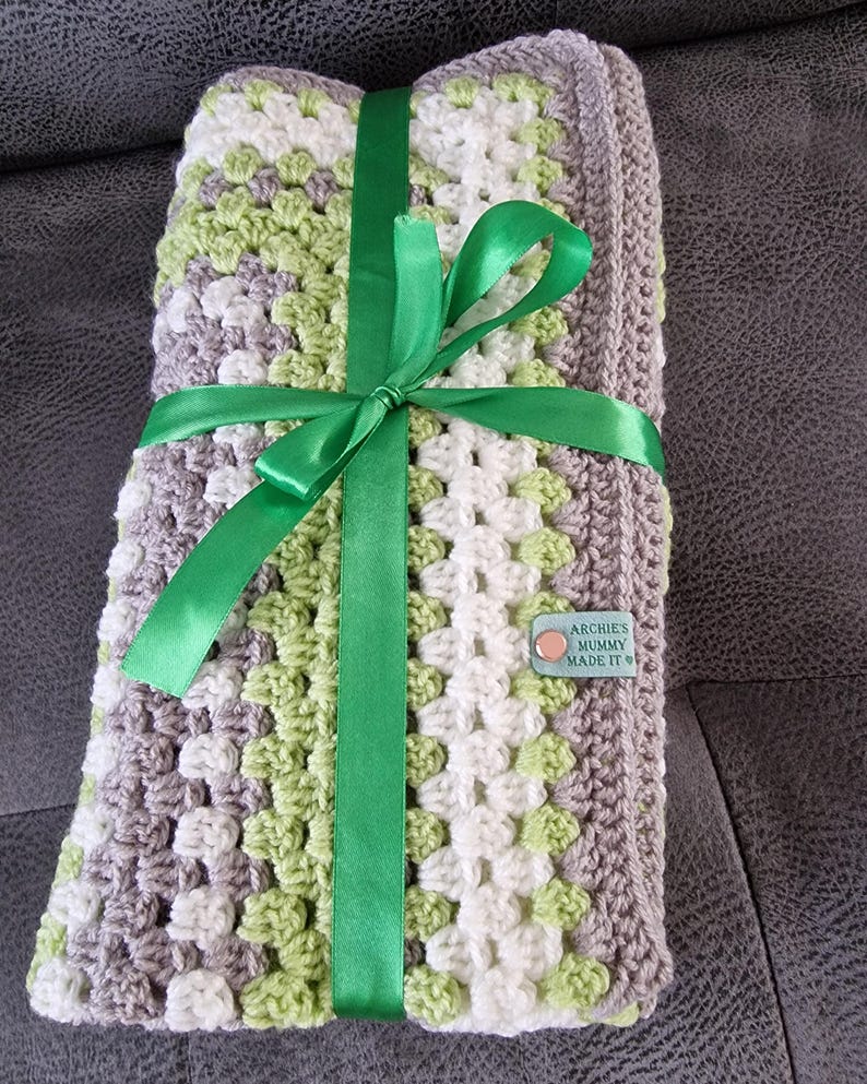 May include: A crocheted baby blanket in shades of gray, white, and green, tied with a green satin ribbon. A small brown tag with the text "Archie's Mummy Made It" is attached to the blanket.