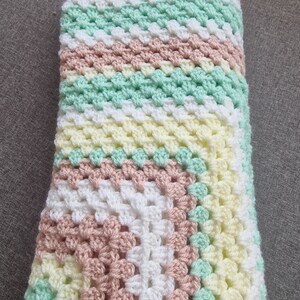 May include: A crocheted baby blanket in shades of pink, white, yellow, and green. The blanket is made with a granny square pattern.