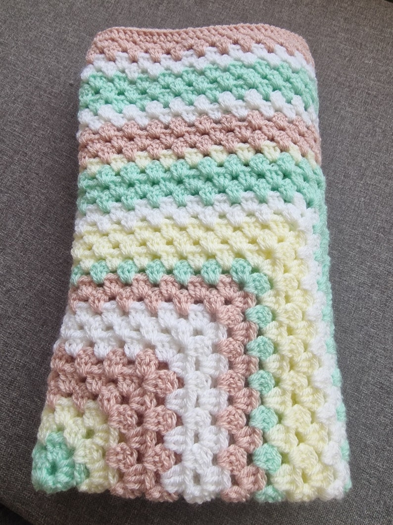 May include: A crocheted baby blanket in shades of pink, white, yellow, and green. The blanket is made with a granny square pattern.