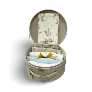 May include: A gray, round baby gift set with a handle, containing a milestone card, swaddling blankets, and a gold ribbon. The milestone card reads "Baby Boy's Milestone Cards". The set includes a light blue and white blanket.
