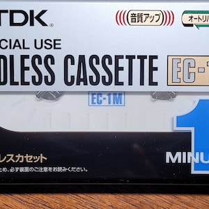 Puede incluir: Paquete de un casete TDK Endless EC-1M. La parte superior plateada tiene el logotipo y texto de TDK. El casete es blanco con un borde negro. El texto "1 MINUTE" está en azul y blanco.