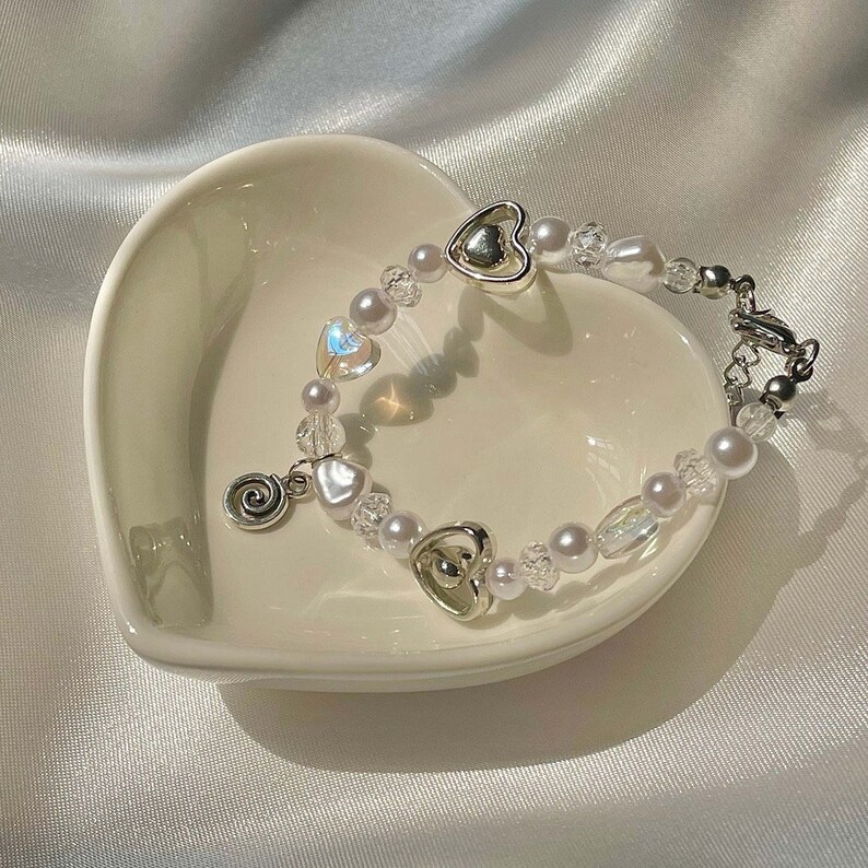 May include: A delicate bracelet featuring a mix of white faux pearls, iridescent beads, and silver heart charms. The bracelet is displayed on a white heart-shaped dish, creating a romantic and elegant aesthetic.