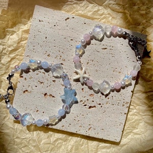 May include: Two beaded bracelets with a sea-themed design. One bracelet features blue star-shaped beads, iridescent shell beads, and white pearls. The other bracelet has pink beads, a white starfish, and iridescent shell beads. Both bracelets have silver chain clasps.