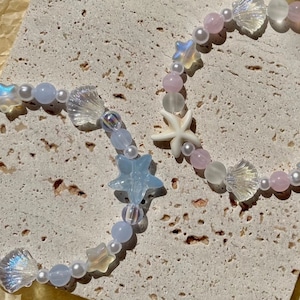 May include: Two beaded bracelets with a sea-themed design. The bracelets feature iridescent star and seashell charms, light blue and pink beads, and small white pearls. A white starfish charm is also included.