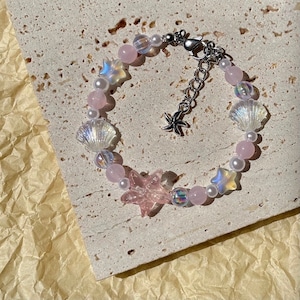 May include: A beaded bracelet featuring pink and iridescent beads, pearls, and seashell and starfish charms. The bracelet has a silver-toned clasp and a small starfish charm at the end of the chain.