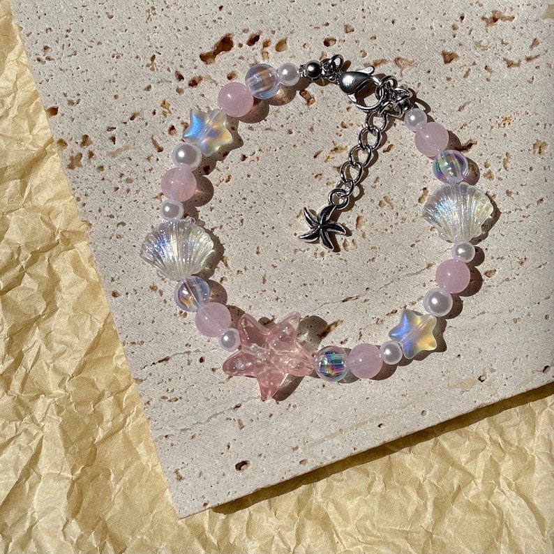 May include: A beaded bracelet featuring pink and iridescent beads, pearls, and seashell and starfish charms. The bracelet has a silver-toned clasp and a small starfish charm at the end of the chain.