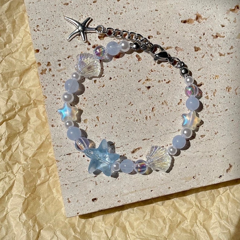May include: A delicate bracelet featuring a starfish charm, iridescent star and shell beads, and light blue and white beads. The bracelet is connected by a silver chain and clasp, creating a coastal theme.