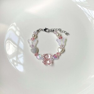 May include: A beaded bracelet featuring a pink teddy bear charm, clear butterfly charms, and heart-shaped beads. The bracelet also includes faux pearls and a silver-toned clasp. The bracelet is designed with a mix of pink, clear, and iridescent beads.