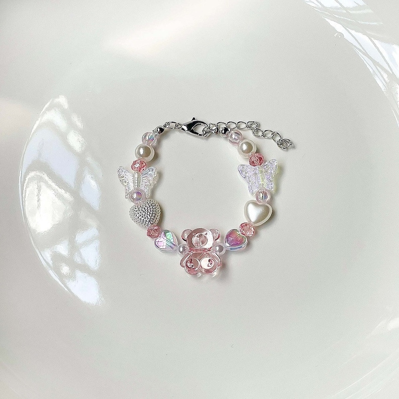 May include: A beaded bracelet featuring a pink teddy bear charm, clear butterfly charms, and heart-shaped beads. The bracelet also includes faux pearls and a silver-toned clasp. The bracelet is designed with a mix of pink, clear, and iridescent beads.