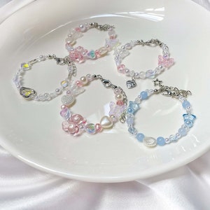 May include: Five beaded bracelets with various designs. The bracelets feature pearls, iridescent beads, and charms in pastel pink, blue, and clear colors. Some bracelets have butterfly, heart, and star charms.