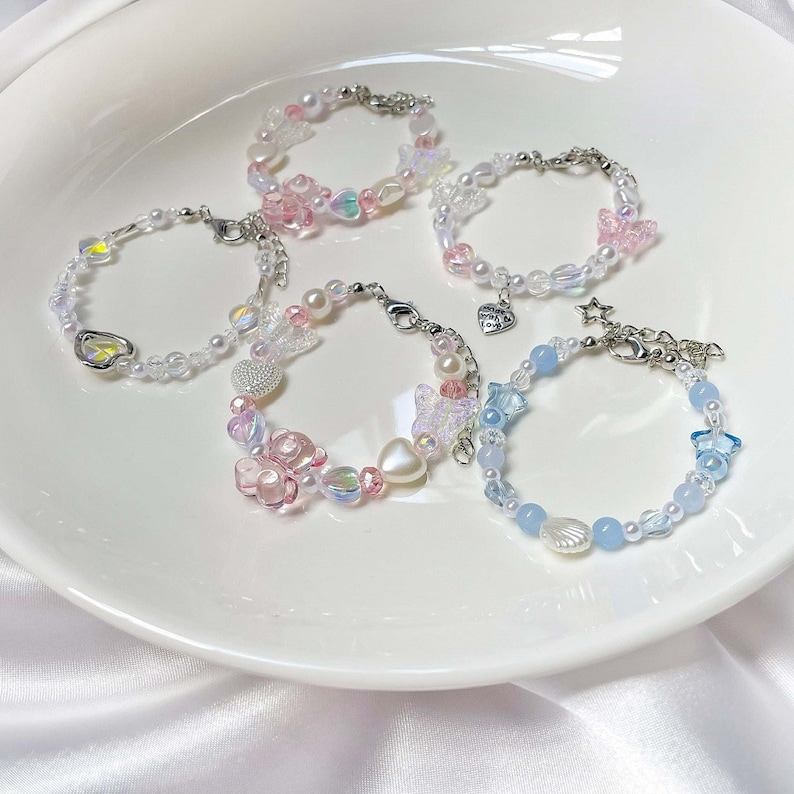 May include: Five beaded bracelets with various designs. The bracelets feature pearls, iridescent beads, and charms in pastel pink, blue, and clear colors. Some bracelets have butterfly, heart, and star charms.