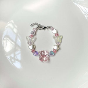 May include: A beaded bracelet featuring a variety of charms, including a pink teddy bear, hearts, stars, and butterflies. The bracelet is made with pink and iridescent beads and has a silver-colored clasp.