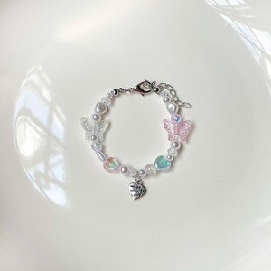 May include: A beaded bracelet featuring iridescent butterfly and heart charms, interspersed with white pearls and clear beads. A small silver heart charm with the words "made with love" hangs from the bracelet. The bracelet has a silver clasp.