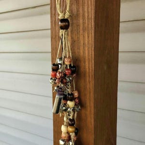 May include: A macrame hanging ornament with wooden beads in various colors, including brown, beige, and red. The ornament also features small silver bells and a rectangular pendant. The natural fiber cord is knotted at the top and bottom.