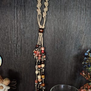 May include: A hanging macrame ornament featuring a knotted jute cord loop at the top, a large dark brown wooden bead, and multiple strands of jute cord adorned with a mix of red, brown, beige, and metallic silver beads and small brass bells.