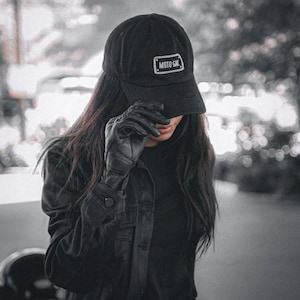 May include: A black moto-style cap with a white embroidered patch that reads "MOTO GAL". The cap is worn by a person wearing a black leather jacket and gloves.