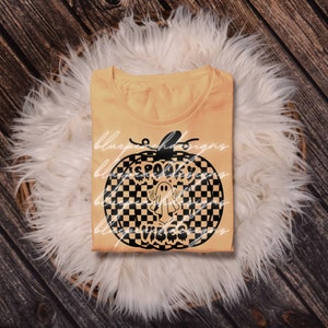 May include: A pale orange t-shirt with a black pumpkin design. The pumpkin graphic features a checkered pattern, the words "Spooky Vibes", and a ghost illustration. The shirt is folded on a white fluffy surface.