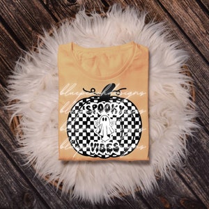 May include: A pale orange t-shirt featuring a black and white chequered pumpkin design. The pumpkin graphic includes a ghost and the words "Spooky Vibes" in white. The shirt is displayed on a white fluffy surface.