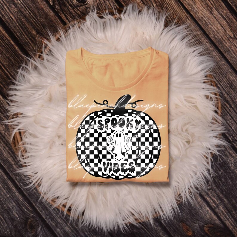 May include: A pale orange t-shirt featuring a black and white chequered pumpkin design. The pumpkin graphic includes a ghost and the words "Spooky Vibes" in white. The shirt is displayed on a white fluffy surface.