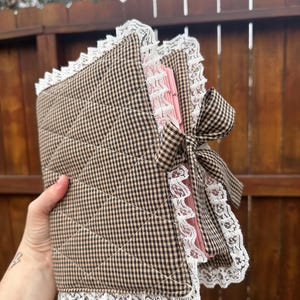 May include: A quilted fabric book cover featuring a brown and black gingham check pattern. The cover is trimmed with white lace along the edges and includes a matching gingham fabric bow tie closure. The interior pages are visible in a soft pink colour.