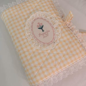 May include: A quilted book cover featuring a yellow and white gingham pattern with white lace trim along the edges. An oval patch in the center displays an embroidered bouquet of pink tulips and the text Proverbs 3:26, framed by lace.