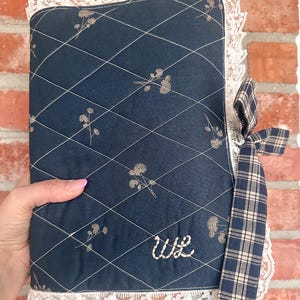 May include: A dark blue quilted fabric book cover featuring a diamond stitch pattern, small floral motifs, and white lace trim along the edges. The cover includes a navy and beige plaid ribbon tie and the embroidered white letters WL on the front.