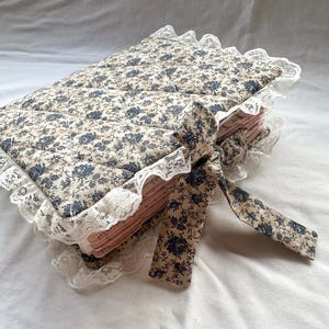 May include: A quilted fabric book cover featuring a beige background with a dark blue floral pattern. The cover is edged with white lace trim and includes a matching fabric tie closure. The pages inside are visible and have a light pink hue.