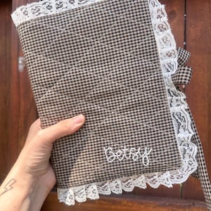 Könnte beinhalten: Ein gesteppter Buchumschlag aus Stoff mit braun-schwarzem Vichy-Karo-Muster. Der Umschlag ist an den Rändern mit weißer Blumen-Spitze verziert und verfügt über ein passendes kariertes Band. Der Name Betsy ist mit weißem Garn eingestickt.