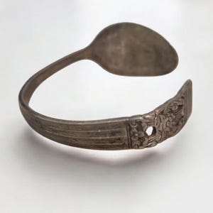 May include: A silver spoon bracelet with a spoon head and a decorative floral design on the cuff. The bracelet is made from a repurposed spoon and has a vintage aesthetic.