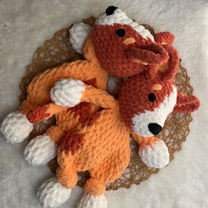 May include: Two crocheted plush toys shaped like dogs, one orange and white and the other red and white, are lying on a woven mat.