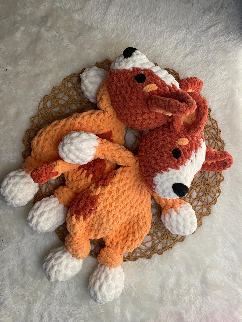 May include: Two crocheted plush toys shaped like dogs, one orange and white and the other red and white, are lying on a woven mat.