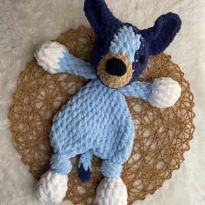 May include: A blue and white crocheted stuffed animal dog toy with a brown nose and black eyes. The toy has white pom-poms for feet.