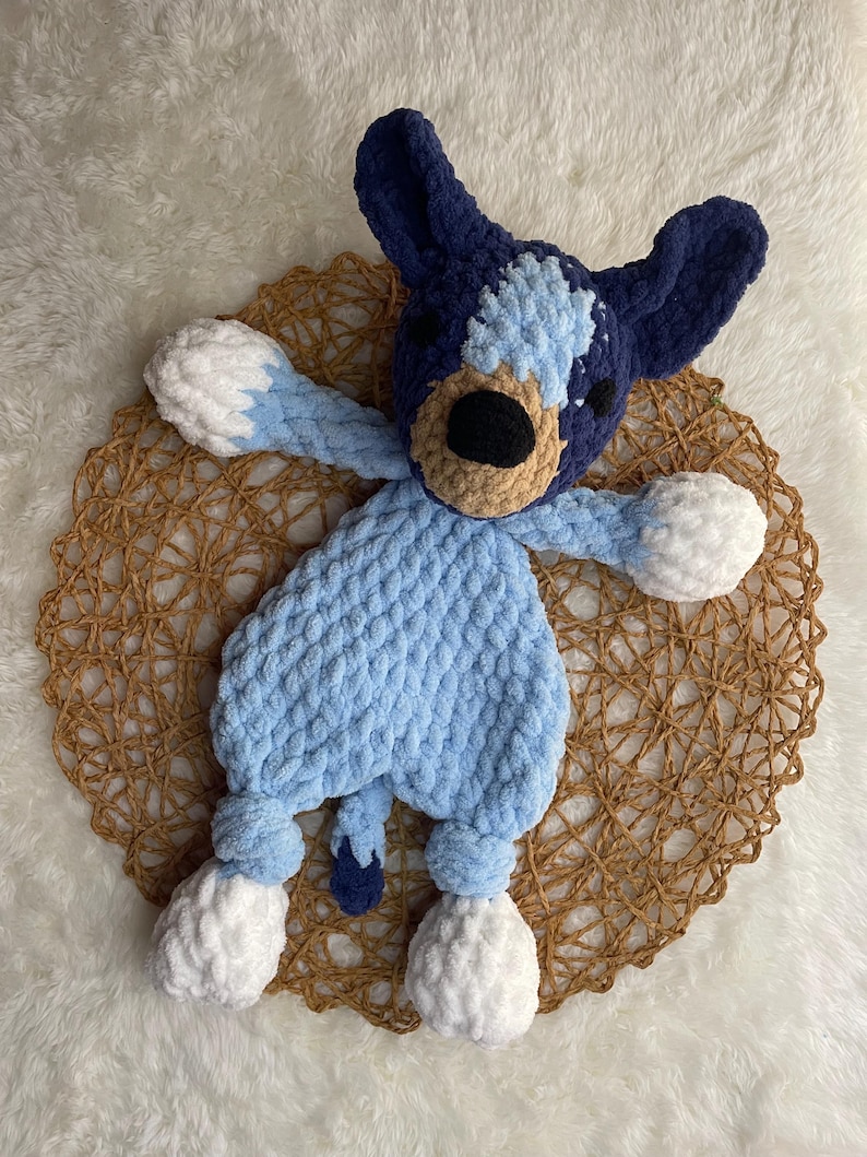 May include: A blue and white crocheted stuffed animal dog toy with a brown nose and black eyes. The toy has white pom-poms for feet.