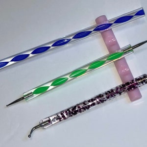 May include: Three nail art tools with clear plastic handles and different coloured designs. The top tool has a blue swirl design, the middle tool has a green swirl design, and the bottom tool has a purple glitter design.
