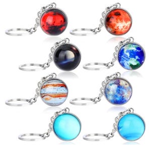 Könnte beinhalten: Acht silberne Schlüsselanhänger mit verschiedenen Planetenmotiven. Jeder Schlüsselanhänger hat einen runden Glasanhänger mit einem anderen Planetenbild, darunter Mars, Jupiter, Erde und Neptun. Die Schlüsselanhänger sind perfekt für Weltraumbegeisterte und Astronomieliebhaber.
