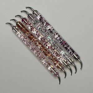 Op de afbeelding: Set van zes nagelkunstgereedschappen met heldere handvatten en metallic glitteraccenten. De gereedschappen hebben een gebogen punt en zijn ontworpen voor precisie nagelkunst.