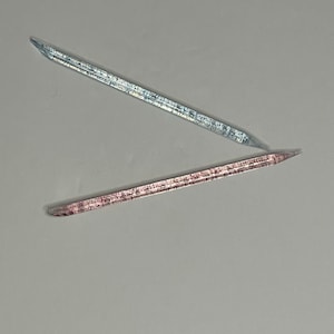 Pode incluir: Duas varinhas de vidro finas e transparentes, uma com um tom azul e a outra com um tom rosa.
