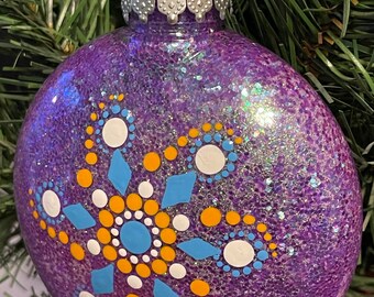 May include: A purple Christmas ornament with a glittery finish and a snowflake design. The snowflake features blue, white, and yellow dot patterns. The ornament hangs from a silver metal cap and a silver string.