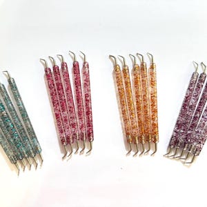 May include: An assortment of glitter-filled nail art tools in shades of teal, pink, orange, and purple. Each tool has a metal tip and a clear, glitter-filled handle. The tools are arranged on a white surface.