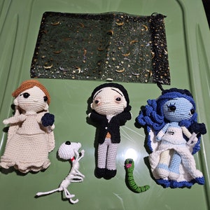 May include: Crocheted amigurumi dolls of various characters, including a bride, a groom, a ghost, and a skeleton. The dolls are made with white, blue, and black yarn. The background is a green and black patterned fabric.