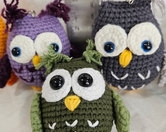 May include: Three crocheted owl toys. The owls are purple, gray, and green. Each owl has large white eyes with black pupils and a yellow beak. The owls are all about 4 inches tall.