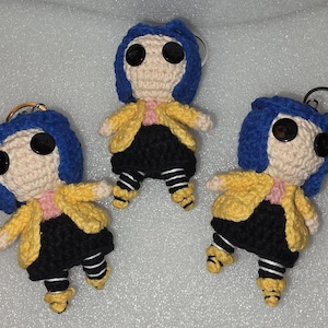 Puede incluir: Tres mu&ntilde;ecas de ganchillo hechas a mano con ojos de bot&oacute;n, pelo azul, chaquetas amarillas, faldas negras y leggings a rayas. Cada mu&ntilde;eca tiene un llavero adjunto. Las mu&ntilde;ecas est&aacute;n de pie, con los brazos extendidos.