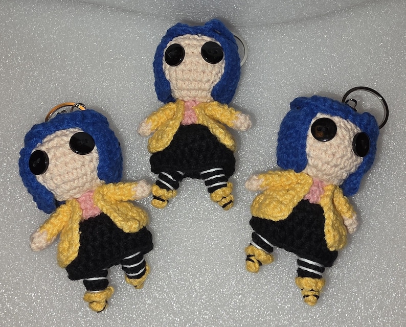 Puede incluir: Tres mu&ntilde;ecas de ganchillo hechas a mano con ojos de bot&oacute;n, pelo azul, chaquetas amarillas, faldas negras y leggings a rayas. Cada mu&ntilde;eca tiene un llavero adjunto. Las mu&ntilde;ecas est&aacute;n de pie, con los brazos extendidos.