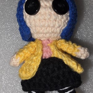 Puede incluir: Mu&ntilde;eca de ganchillo con una cabeza beige claro, grandes ojos negros de bot&oacute;n y cabello azul. La mu&ntilde;eca viste una chaqueta amarilla, una camisa rosa, una falda negra y mallas a rayas blancas y negras. Los zapatos son amarillos.