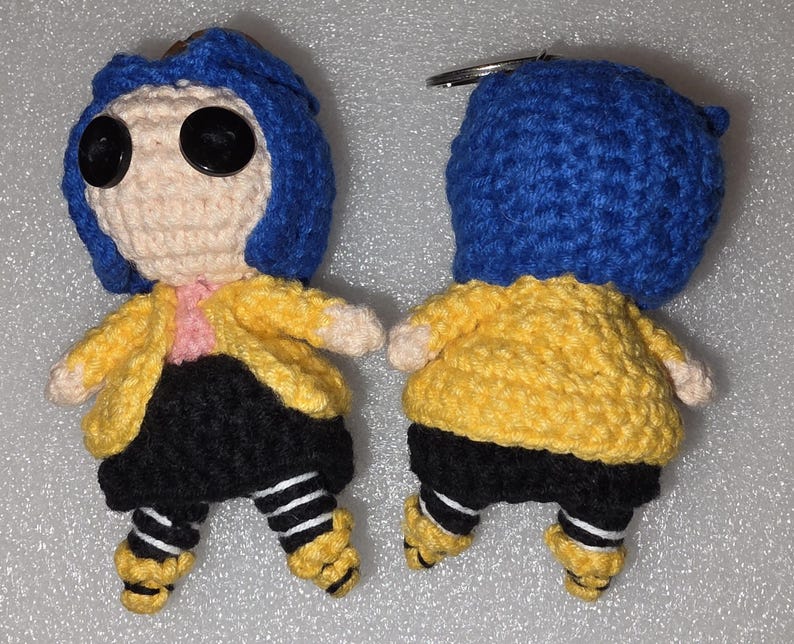 Puede incluir: Mu&ntilde;eca de ganchillo con cabello azul, ojos de bot&oacute;n y una chaqueta amarilla. La mu&ntilde;eca tiene pantalones cortos negros, calcetines a rayas y zapatos amarillos. Tiene un llavero adjunto.
