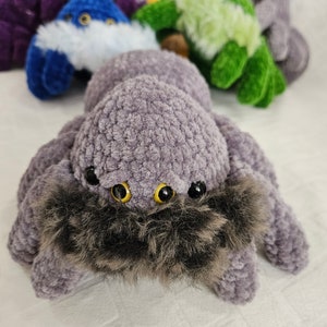 May include: A plush purple spider toy with black fur around its face and yellow eyes. The spider is lying on a white surface.