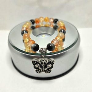 May include: A beaded stretch bracelet featuring translucent orange and yellow crackle glass beads interspersed with smooth black beads. A silver-toned metal charm shaped like a butterfly with skull details on its wings hangs from the center.
