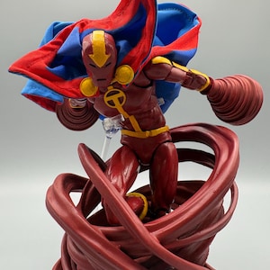 May include: A posable action figure featuring a maroon and yellow suit with a matching helmet. The figure wears a vibrant red and blue fabric cape and is positioned atop a swirling, sculpted maroon base that mimics a tornado or wind effect.